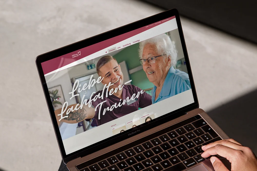 Laptopbildschirm zeigt eine Website mit einer lächelnden Pflegekraft neben einer älteren Frau und der Überschrift "Liebe, Lachen, Trauern", offenbar für Seniorenbetreuung oder Pflegeangebote.