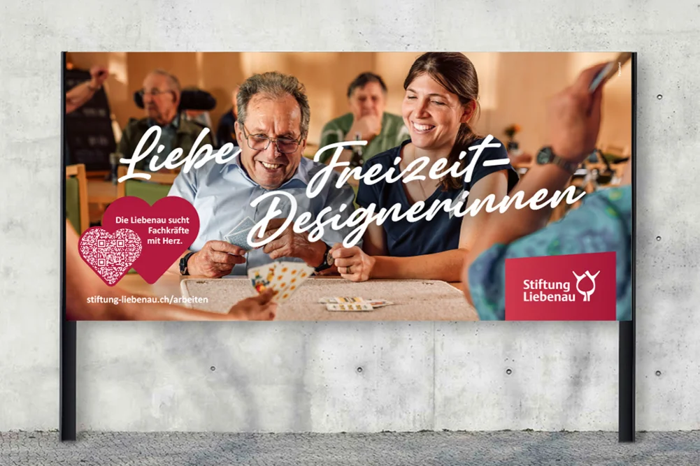 Werbeplakat: Eine lächelnde junge Frau und ein älterer Mann spielen Karten in einer Gruppe; freundliche Betreuungsszene mit Schriftzug Liebe Freizeit-Designerinnen und Logo der Stiftung Liebenau.