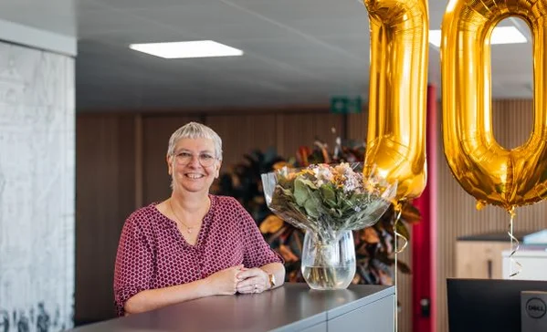 Lächelnde Frau mit kurzem grauem Haar steht hinter einem Empfangstresen, neben einer Vase mit Blumenstrauß und großen goldenen Zahlenballons.