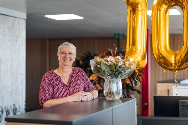 Lächelnde Frau mit kurzem grauem Haar steht hinter einem Empfangstresen, neben einer Vase mit Blumenstrauß und großen goldenen Zahlenballons.