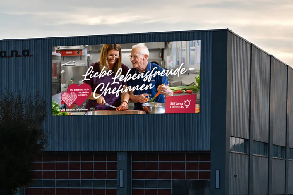 Außenwerbung mit Foto eines älteren Mannes und einer jüngeren Frau, die gemeinsam lächelnd kochen, dazu der Slogan Liebe Lebensfreude-Chefinnen und das Stiftung Liebenau-Logo.