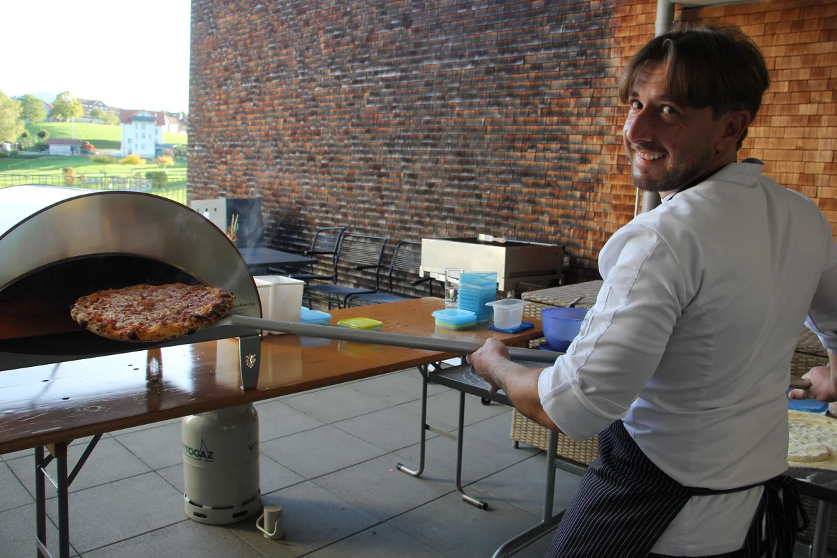 Koch in weißer Jacke lächelt zur Kamera, während er mit einer langen Schaufel eine frisch gebackene Pizza aus einem tragbaren Holzofen auf einer Terrasse holt