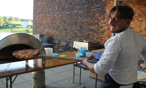 Koch in weißer Jacke lächelt zur Kamera, während er mit einer langen Schaufel eine frisch gebackene Pizza aus einem tragbaren Holzofen auf einer Terrasse holt