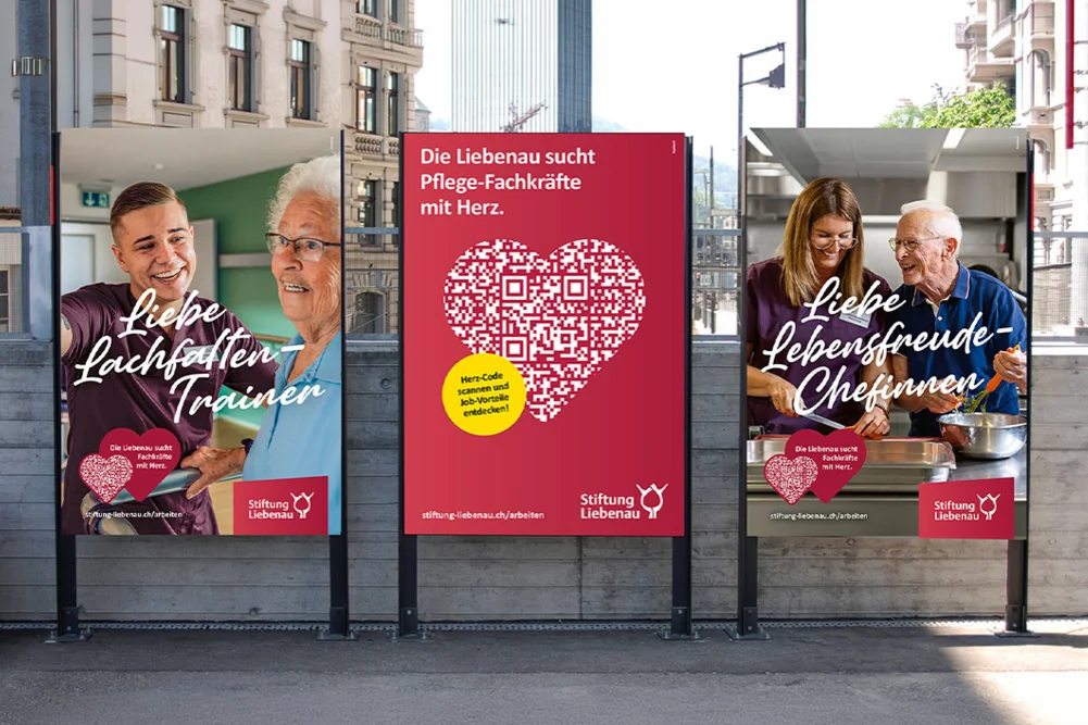 Drei bunte Werbeplakate einer Pflegestiftung an einer Haltestelle: mittiges pinkes Plakat mit QR-Code in Herzform und Text Die Liebenau sucht Pflege-Fachkräfte mit Herz, links und rechts Fotos lachender Pflegerinnen und Senioren, Motiv warme Betreuung und Einladung zur Bewerbung.